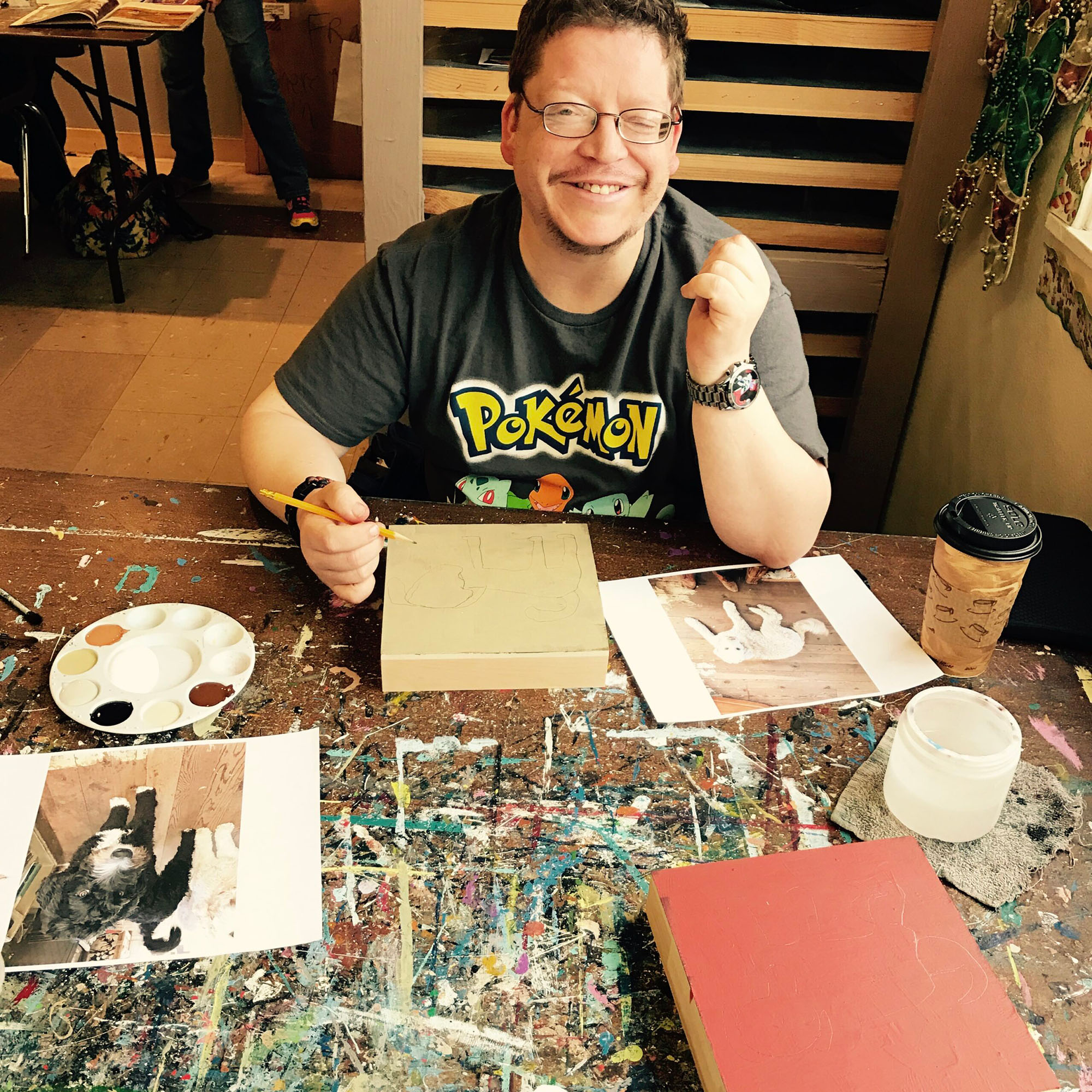 A smiling person preparing to paint a picture. There are several reference photos of dogs on the table