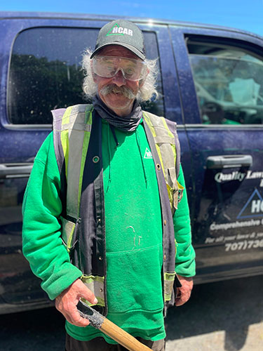 A man in working clothes stands smiling in front of a truck while holding a shovel