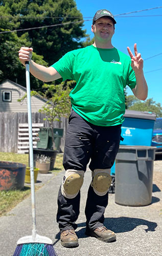 A smiling man in working clothes stands on a sidewalk while holding a broom