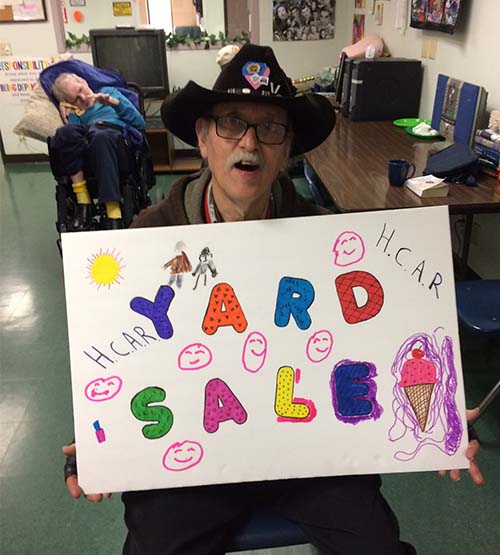 A happy man holds a large, colorful yard sale sign. In the background is another person in a wheelchair.