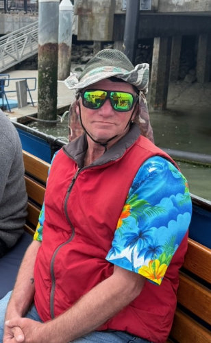 A person in sunglasses, a vest, and a vibrant Hawaiian-style shirt sits on a bench